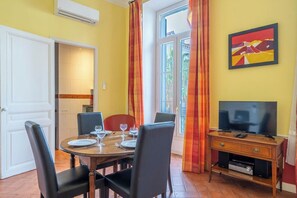 A dining area with a wooden table set for four, chairs, a television on a wooden stand, and a window with curtains. - Le 10 Jean Daumas avec 1 Chambre et Balcon (Cannes)