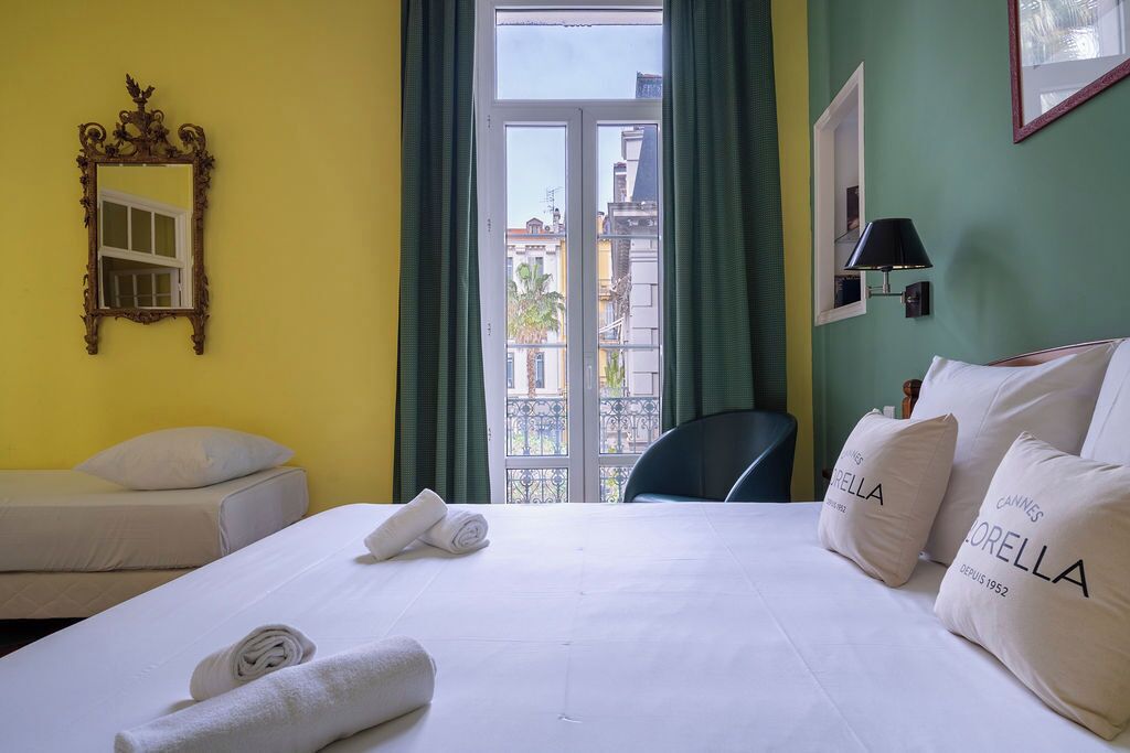 Ein Hotelzimmer mit einem Bett, zwei Nachttischen, einem Spiegel, einem Fenster mit Vorhängen und Blick auf Gebäude.