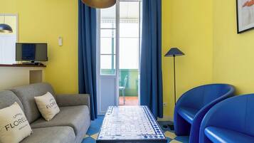 Un salon avec un canapé, une table et deux fauteuils bleus. On y trouve également un téléviseur posé sur un meuble et une lampe.