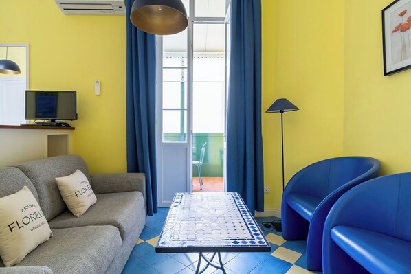A living room with a sofa, a table, and two blue chairs. There is a television on a stand and a lamp. - Le 10 Jean Daumas Studio avec Loggia (Cannes)