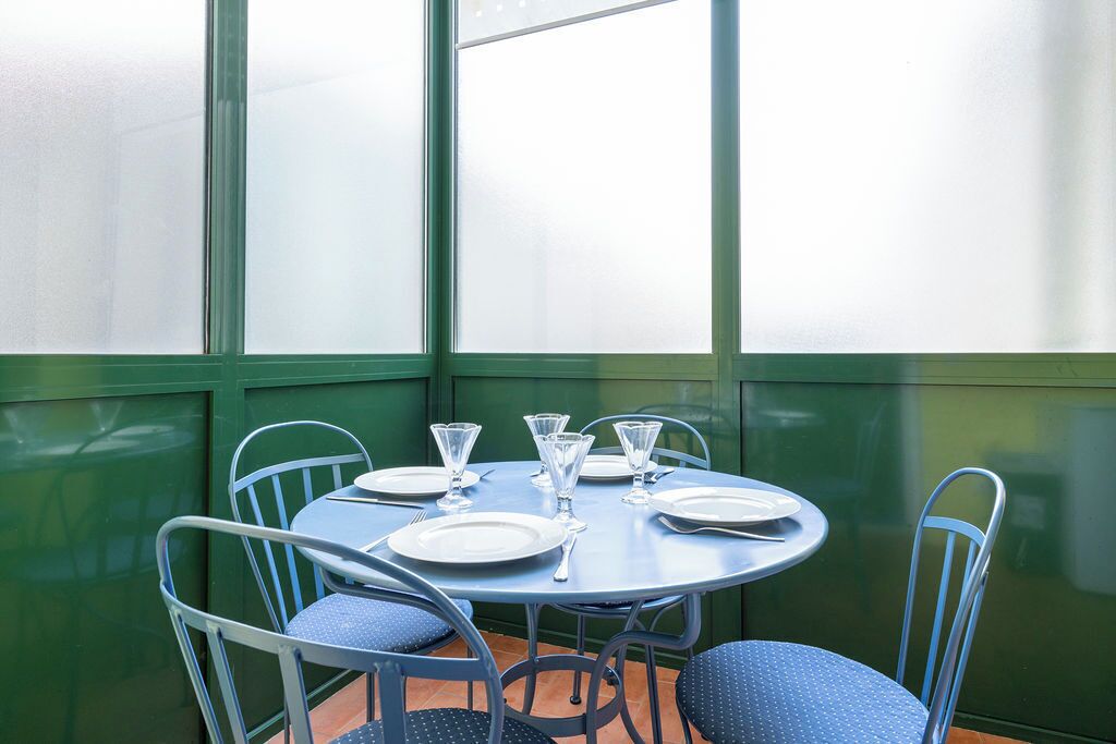 Une table ronde, dressée avec des assiettes, des couverts et des verres, entourée de chaises en métal dotées de coussins bleus, dans une pièce aux murs verts et aux grandes fenêtres.
