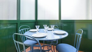 Taula rodona coberta amb plats, coberts i gots, envoltada de cadires metàl·liques amb coixins blaus, en una habitació amb parets verdes i grans finestres.