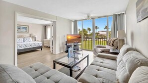 A hotel room with a bed, a sofa, a television, and a balcony with a view of palm trees.