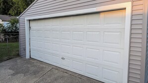 Une porte de garage blanche avec une poignée en métal, située dans un quartier résidentiel, délimitée par une clôture en treillis métallique et donnant sur une pelouse.