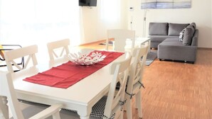 Un área de comedor con una mesa blanca, un mantel individual rojo y sillas blancas. Un sofá gris se ve al fondo.