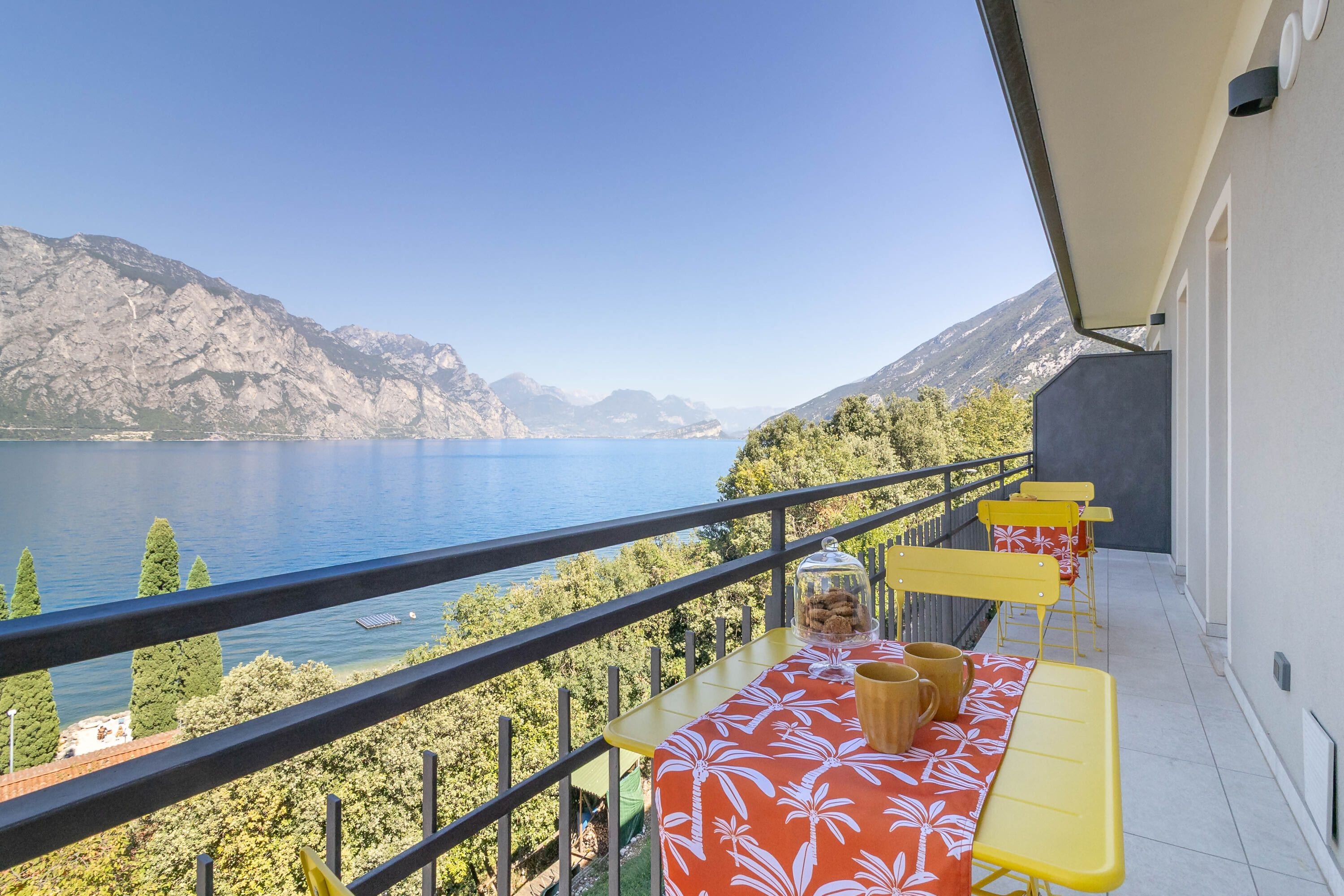 A balcony with a view of a lake and mountains, yellow and red furniture, and a clear sky.