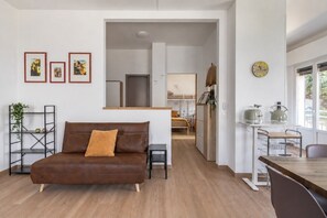 Un salon moderne avec un canapé en cuir marron, une petite table et un coin cuisine visible à travers une porte ouverte.