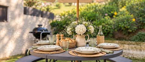 An outdoor dining setup with a round table, multiple plates, glasses, and a central floral arrangement.