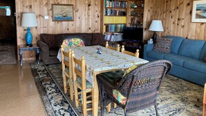 Ein gemütliches Wohnzimmer mit einer holzverkleideten Wand, einem Esstisch mit Stühlen, einem blauen Sofa, einem Bücherregal und einem Fernseher.
