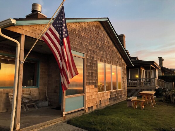 A wooden house with a porch, large windows, and an American flag. - Dot's Beachfront Bungalow Off-Grid Haven (Arch Cape)
