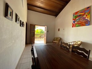 Quarto espaçoso com teto de madeira, mesa de jantar com cadeiras, pintura abstrata colorida e porta que dá para área externa.