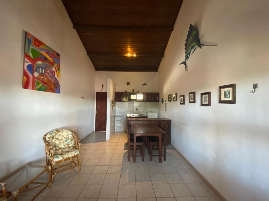 Une pièce comprenant une table à manger, des chaises, un fauteuil en osier, un coin cuisine et un mur orné de tableaux.