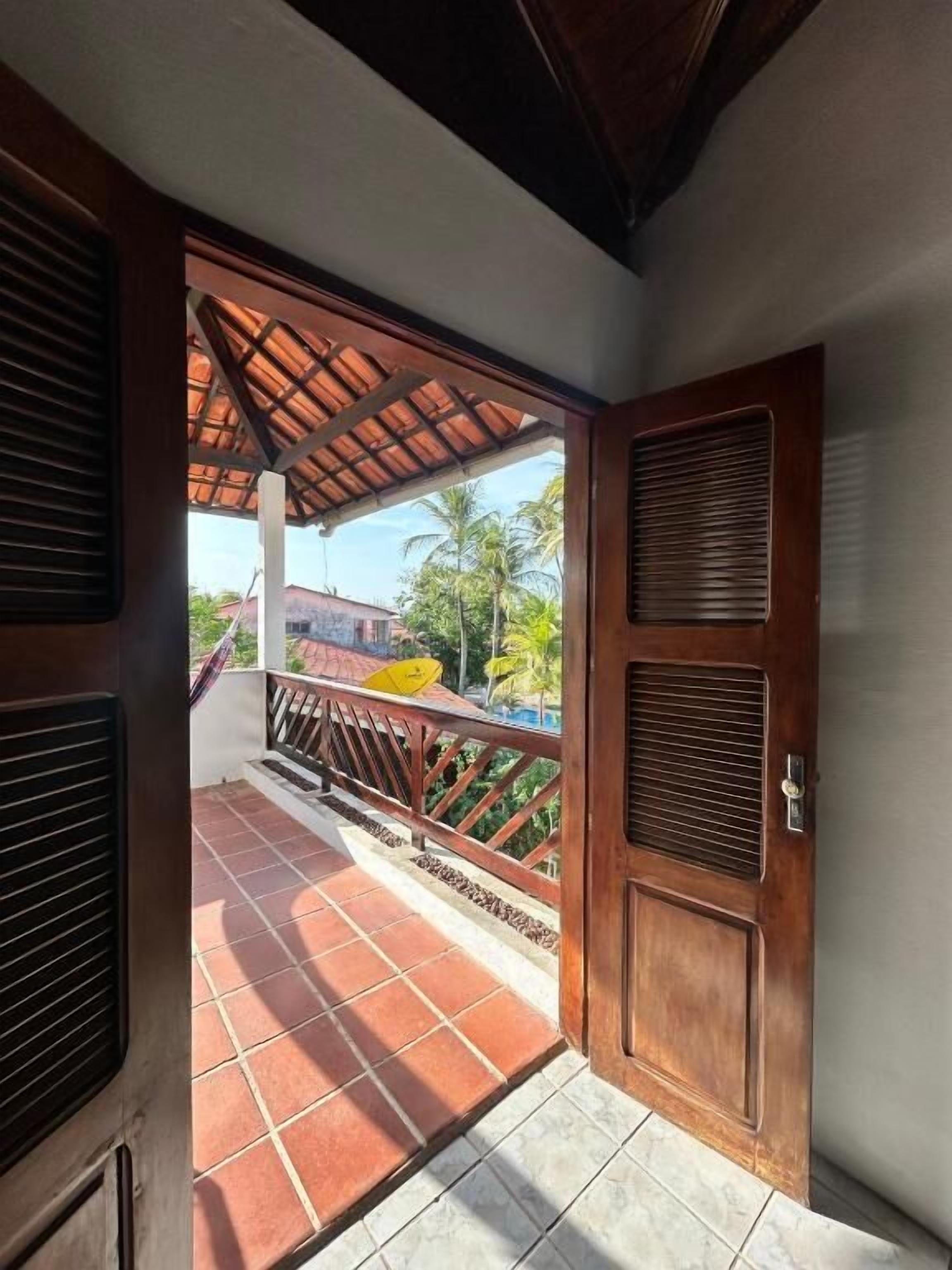 A wooden door leading to a tiled balcony with a view of palm trees and a building.
