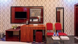 A hotel room with a wooden desk, a TV, a mirror, a red chair, a small table with a tray, and a potted plant. - Gulf Star Hotel (Dubai)