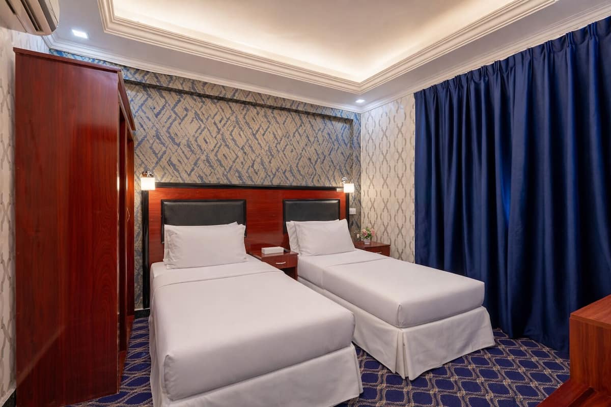 A hotel room with two single beds, a wooden wardrobe, a nightstand with a lamp, and a patterned carpet.