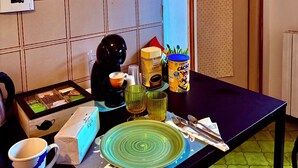 A room with a tiled wall, a television mounted on the wall, a table with a coffee maker, a mug, a plate, and a box. - Vanilla Center-2 Rooms DBL+ Equipped Kitchen (Torino)
