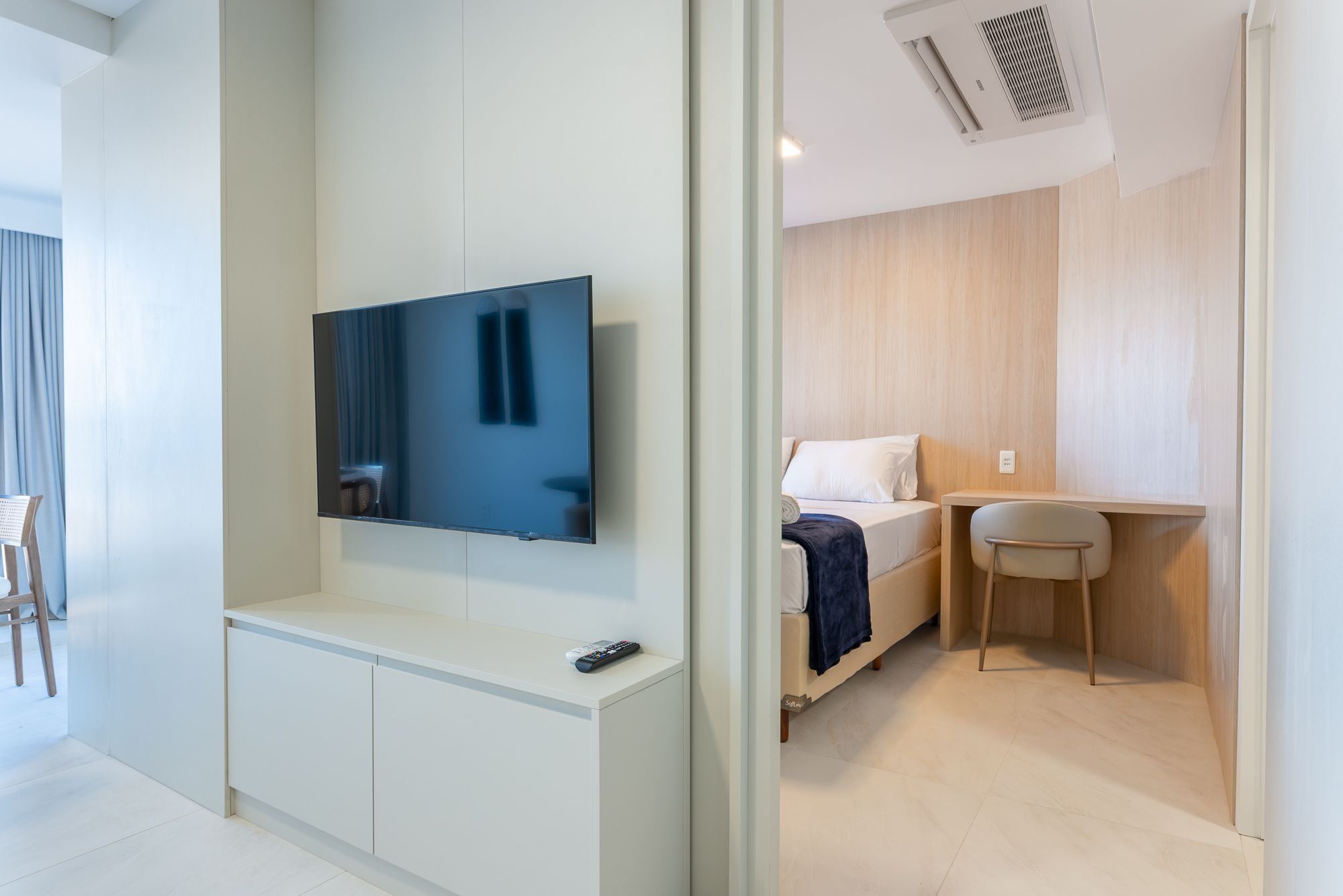 A modern hotel room with a flat-screen TV mounted on the wall, a small desk with a chair, a bed with a blue blanket, and a wooden panel wall.
