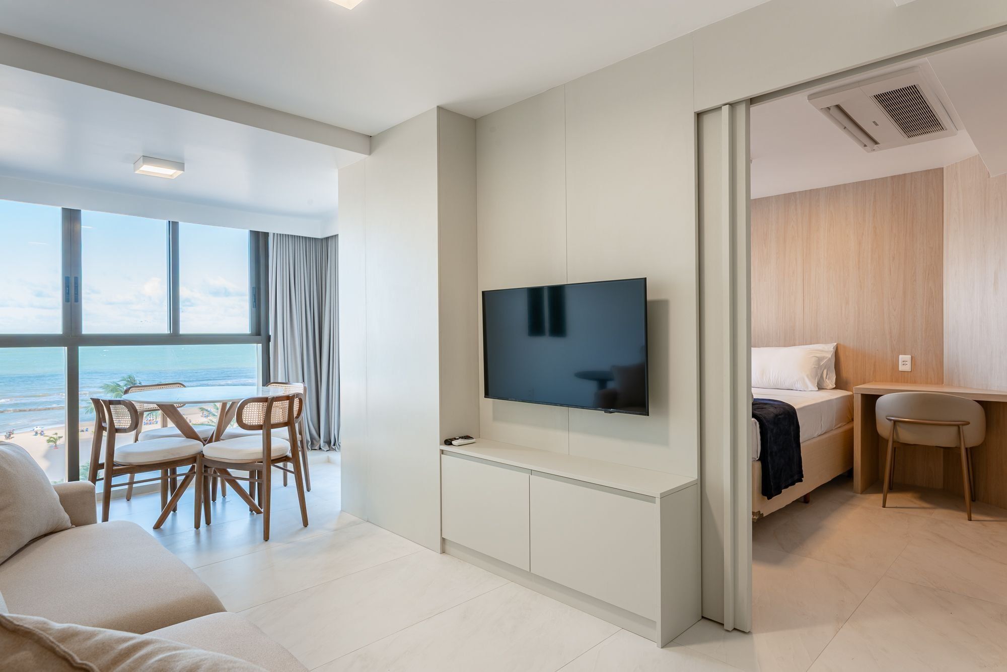 Una habitación de hotel moderna con un gran ventanal que ofrece vistas a la playa, un televisor de pantalla plana montado en la pared, una cama con cabecera de madera y un pequeño escritorio con una silla.