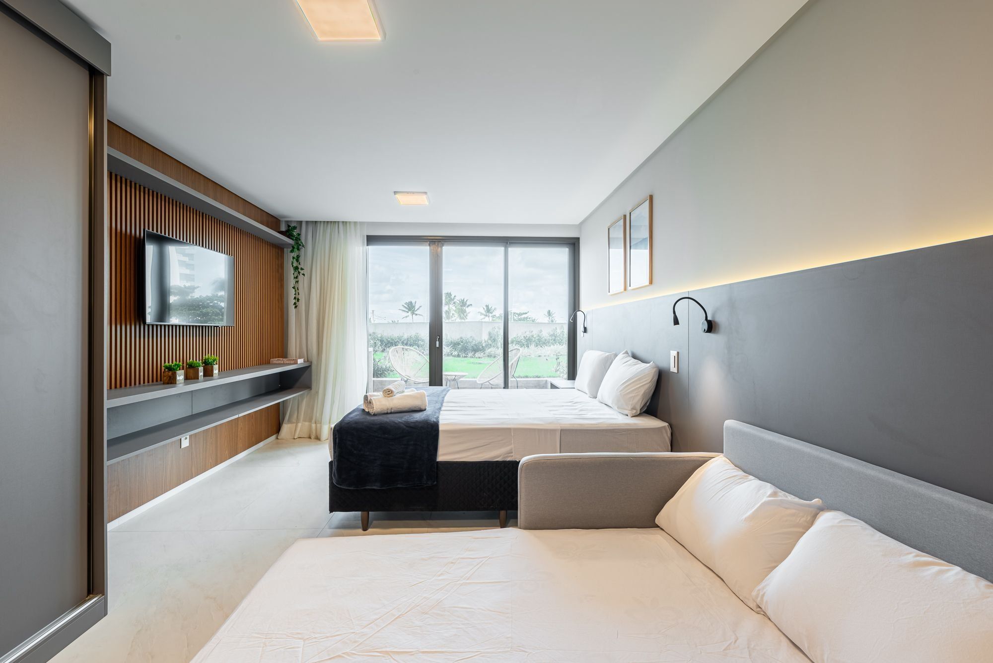 A modern bedroom with a large bed, a smaller bed, a sofa, a TV mounted on a wooden panel, and a large window offering a view of the outdoors.