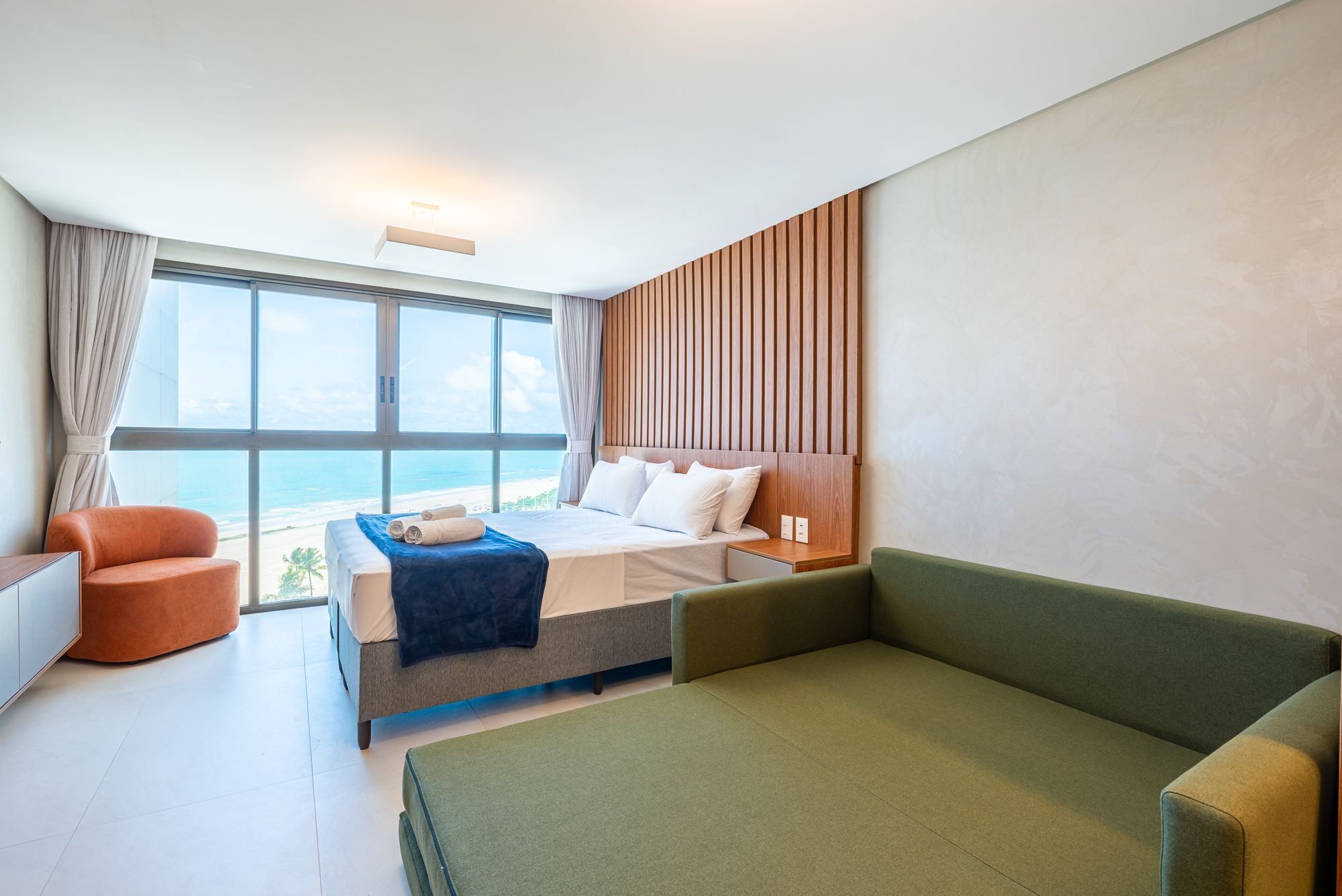 Una habitación de hotel con una cama, un sofá verde, un sillón naranja pequeño y vistas al océano.