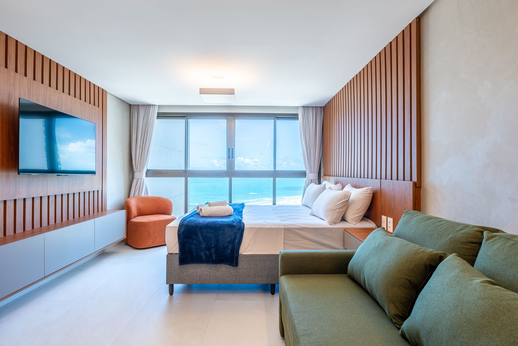 A modern hotel room with a bed, a sofa, a TV, and a view of the sea.