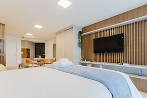 Una habitación de hotel moderna con una cama grande, un televisor de pantalla plana montado en una pared de paneles de madera, un escritorio pequeño con una silla y una zona de comedor al fondo.
