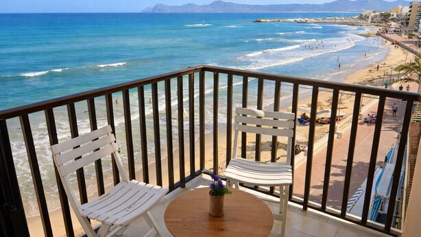 Een balkon met twee witte stoelen en een kleine tafel, met uitzicht op een strand met mensen en een berg op de achtergrond.