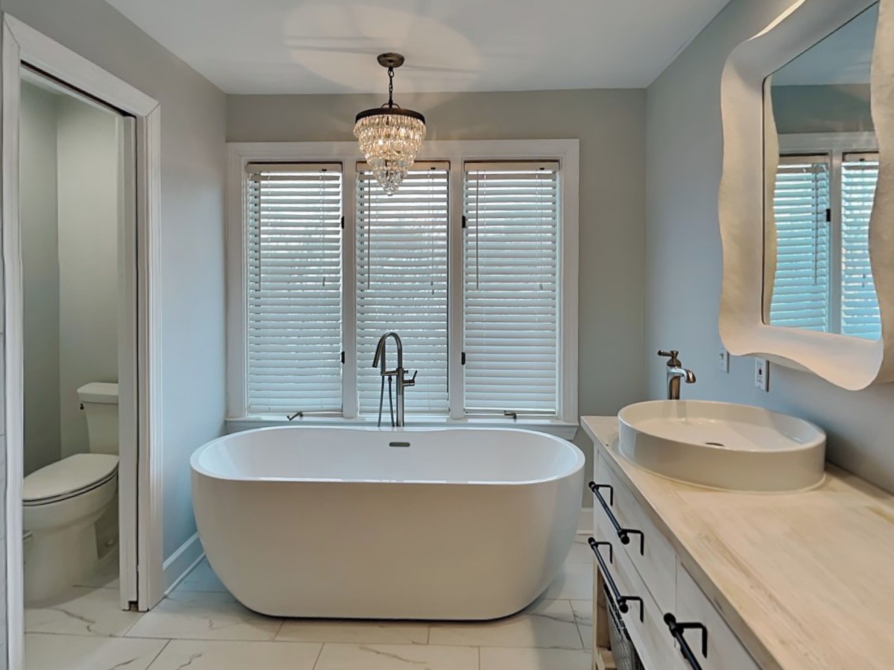 A modern bathroom with a freestanding bathtub, a sink, a chandelier, and a window with blinds.