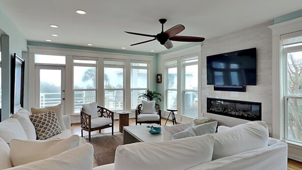 A spacious living room with a ceiling fan, white furniture, large windows, and a fireplace. - The Northshore (Tybee Island)