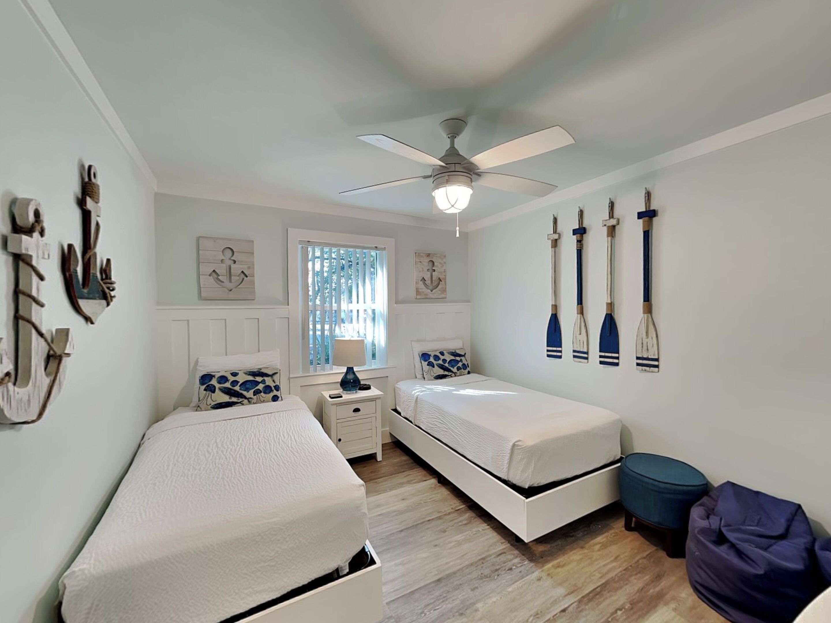 Un dormitorio con dos camas, ventilador de techo, ventana con cortinas y decoración de temática náutica.