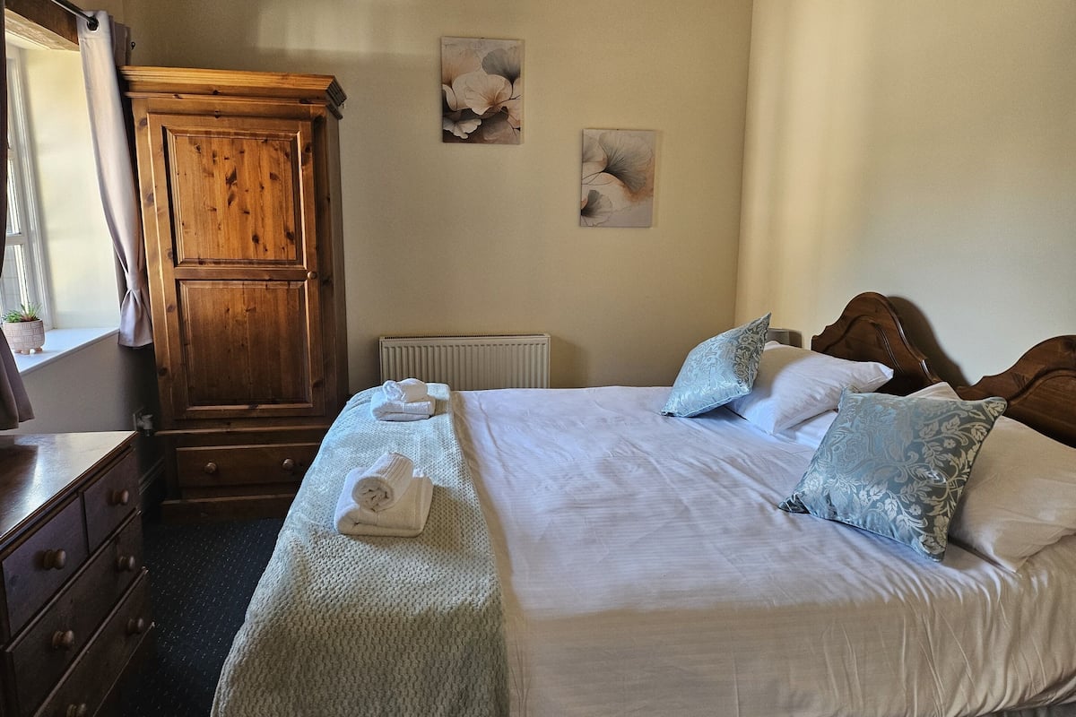 Una camera da letto con un armadio in legno, un letto con biancheria da letto bianca e cuscini fantasia, e due quadri floreali.
