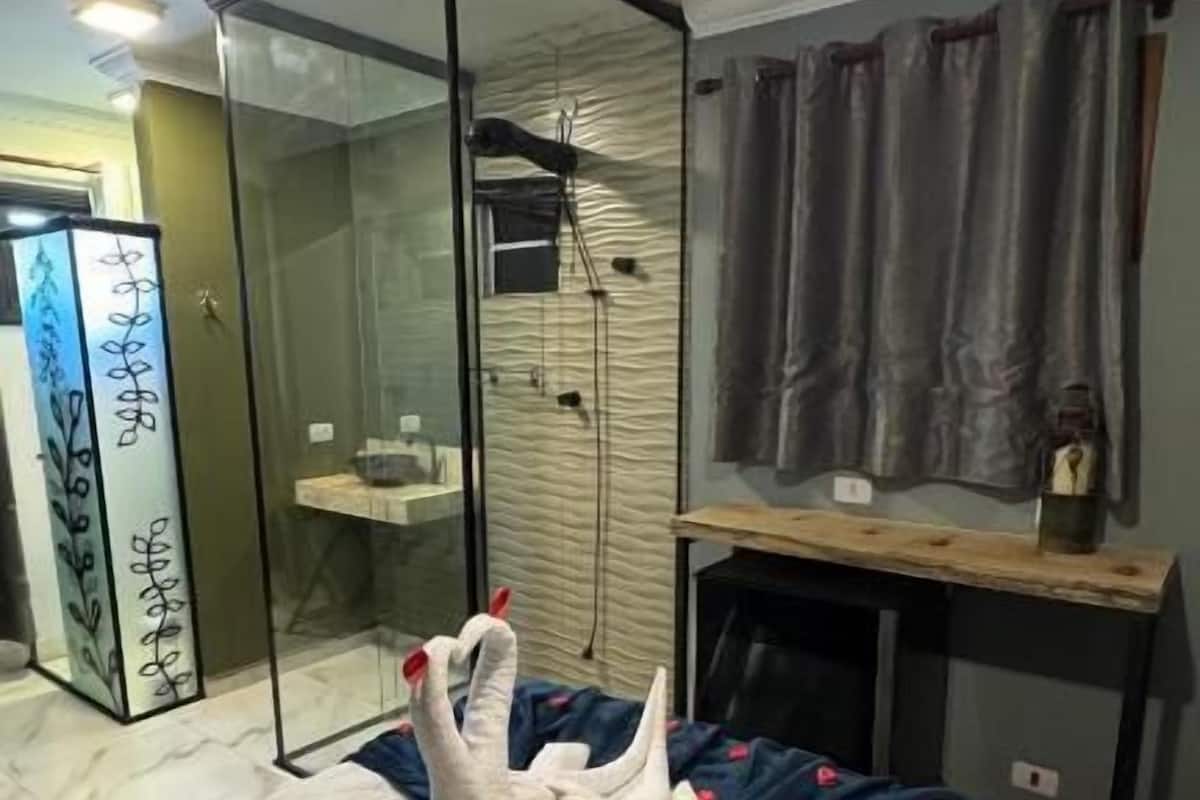 A modern hotel room with a glass-enclosed shower, a bed with a white and blue duvet, and a hanging light fixture.