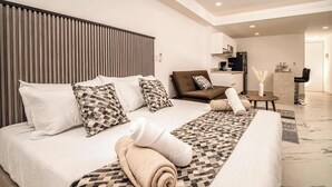 Quarto de hotel moderno com uma cama grande, um sofá, uma mesinha e uma cozinha compacta.