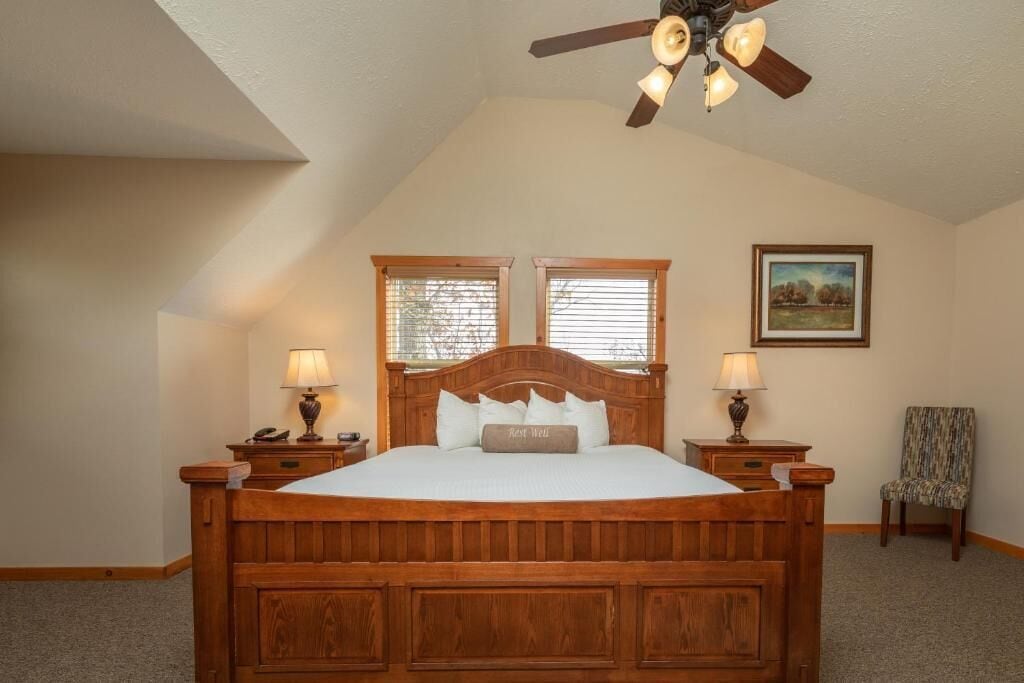 Une chambre à coucher avec un lit en bois, deux lampes de chevet, un ventilateur de plafond, une fenêtre et une chaise.
