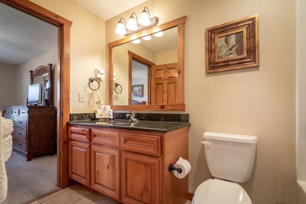 Une salle de bain avec un meuble-lavabo en bois, des toilettes, un miroir avec éclairage intégré et un tableau encadré au mur.