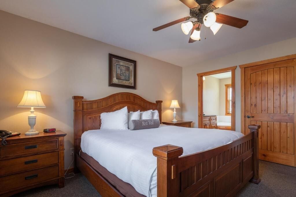 Une chambre à coucher avec un lit en bois, deux tables de chevet avec des lampes, un ventilateur de plafond, un tableau encadré accroché au mur et une porte en bois.
