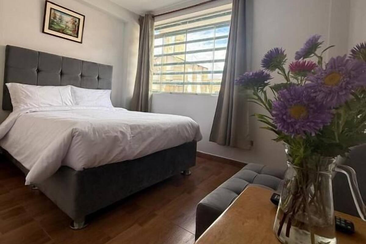 A hotel room with a bed, a TV mounted on the wall, a window with curtains, and a vase with flowers on a table.