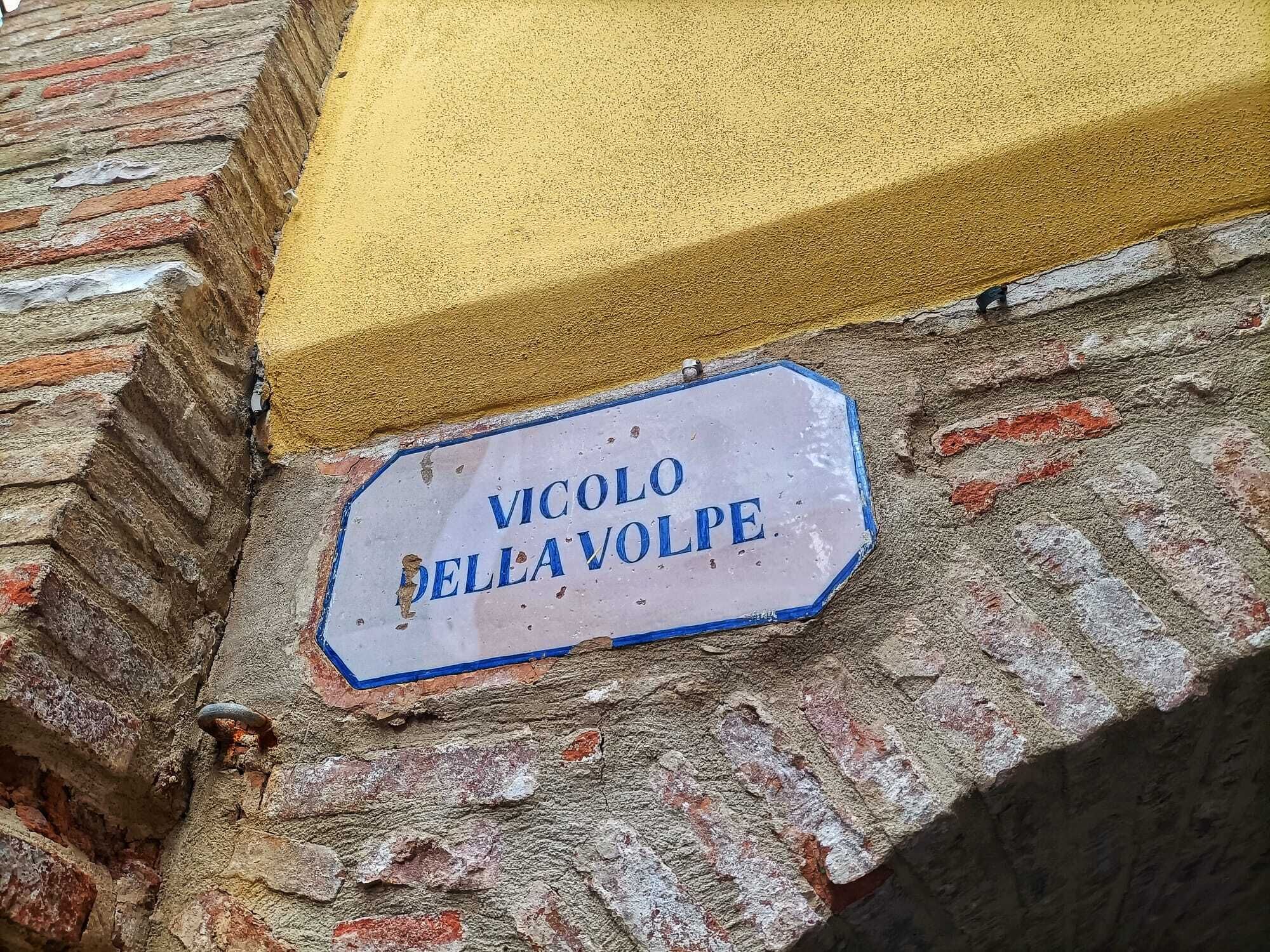 Un letrero desgastado con la inscripción 'VICOLLO DELLA VOLPE', sujeto a una pared de ladrillo con una sustancia amarillenta en la parte superior.