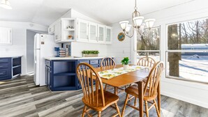 A modern kitchen with white cabinets, a wooden dining table with chairs, and a view of a snowy landscape through large windows. - Feather Trail Guest House - 3BR Cozy Family Stay (Nekoosa)