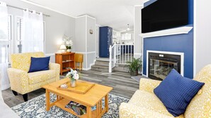 A living room with patterned sofas, a coffee table, a fireplace, and a staircase leading to a balcony. - Feather Trail Guest House - 3BR Cozy Family Stay (Nekoosa)