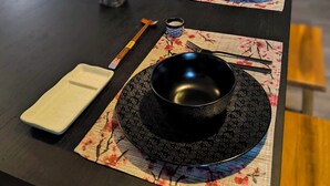 A dining table set with black bowls, chopsticks, a small cup, and a vase with pink flowers. - Zen Sakura - The Breath of Asia! (Belfort)
