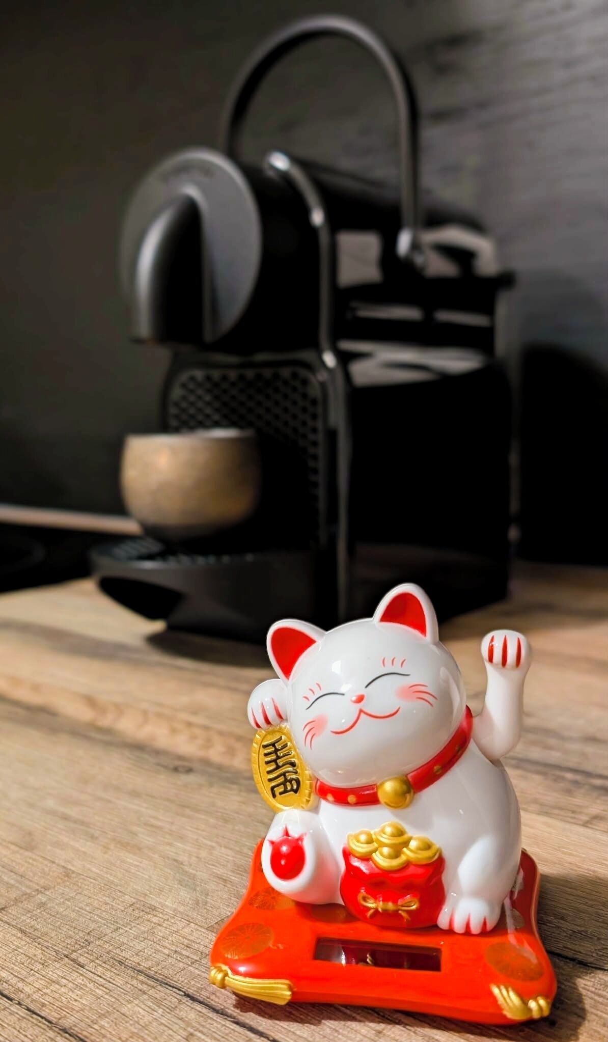 Une statuette de chat blanc, portant un collier rouge et agrémentée de détails dorés, assise sur un socle rouge avec une pièce d’or et une clochette rouge.
