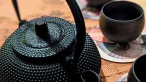 A black teapot with a textured handle and spout, placed on a woven mat with several small cups and saucers nearby. - Zen Sakura - The Breath of Asia! (Belfort)