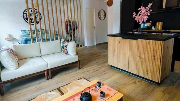 A modern living room with a sofa, a wooden partition, a dining table with tea set, and a kitchen area with a black countertop and wooden cabinets. - Zen Sakura - The Breath of Asia! (Belfort)