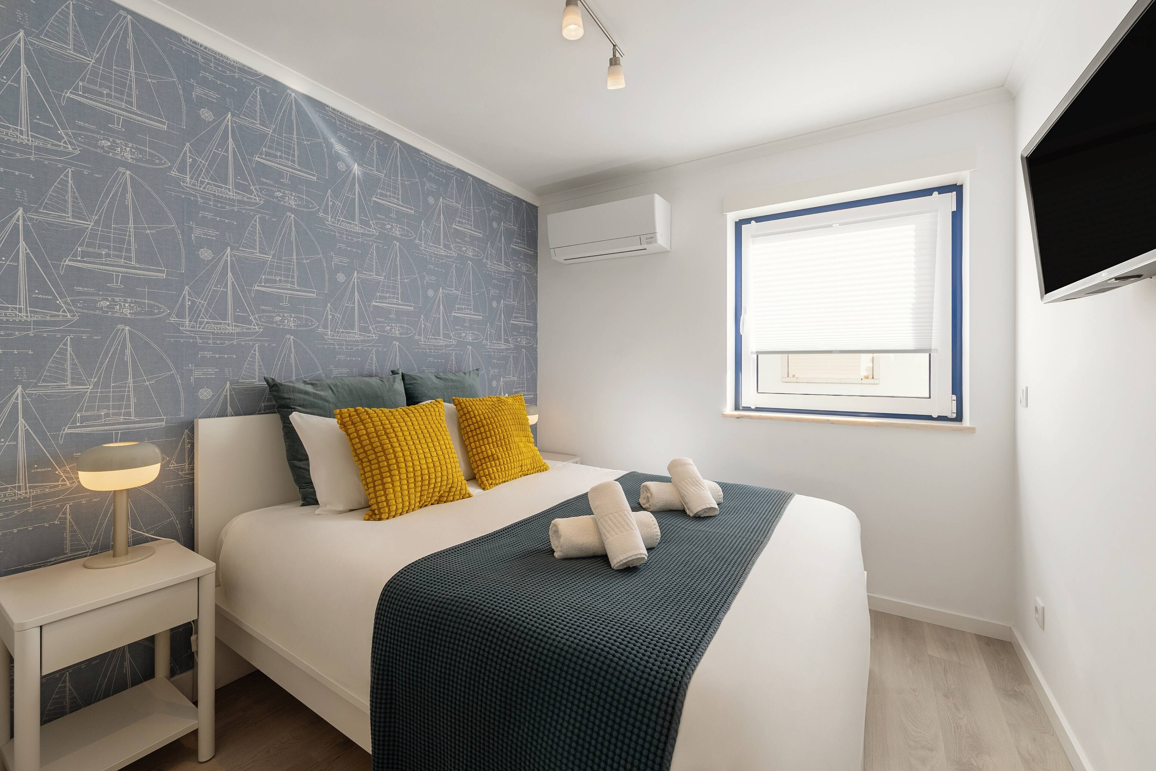 A neatly made bed with a dark blanket, two pillows, and rolled towels. There is a bedside table with a lamp, a window with blinds, and a wall-mounted air conditioner.