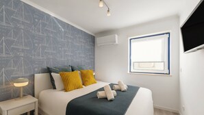 Una cama bien hecha con un edredón oscuro, dos almohadas y toallas enrolladas. Se ve una mesita de noche con una lámpara, una ventana con persianas, y un aire acondicionado instalado en la pared.
