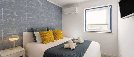 A neatly made bed with a dark blanket, two pillows, and rolled towels. There is a bedside table with a lamp, a window with blinds, and a wall-mounted air conditioner.