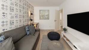 Una sala de estar con un televisor de pantalla plana grande, un sofá gris, cojines estampados, una pared con ilustraciones de vida marina y un cuadro de un velero.