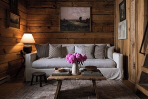 A cozy room with a wooden wall, a white sofa, a small wooden table with books and a vase of flowers, and a lamp. - Asheville Ivy Cabin - Superhost (Black Mountain)