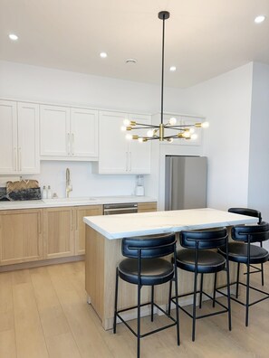 Una cocina moderna con armarios blancos, un refrigerador de acero inoxidable, una encimera de mármol y una zona con taburetes de bar.